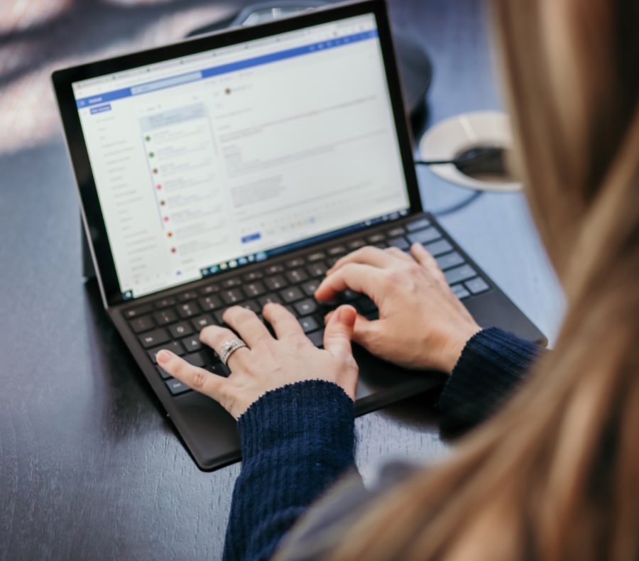 A person in a blue sweater types on a laptop with an email inbox open, seen from behind focusing on their hands and the laptop.