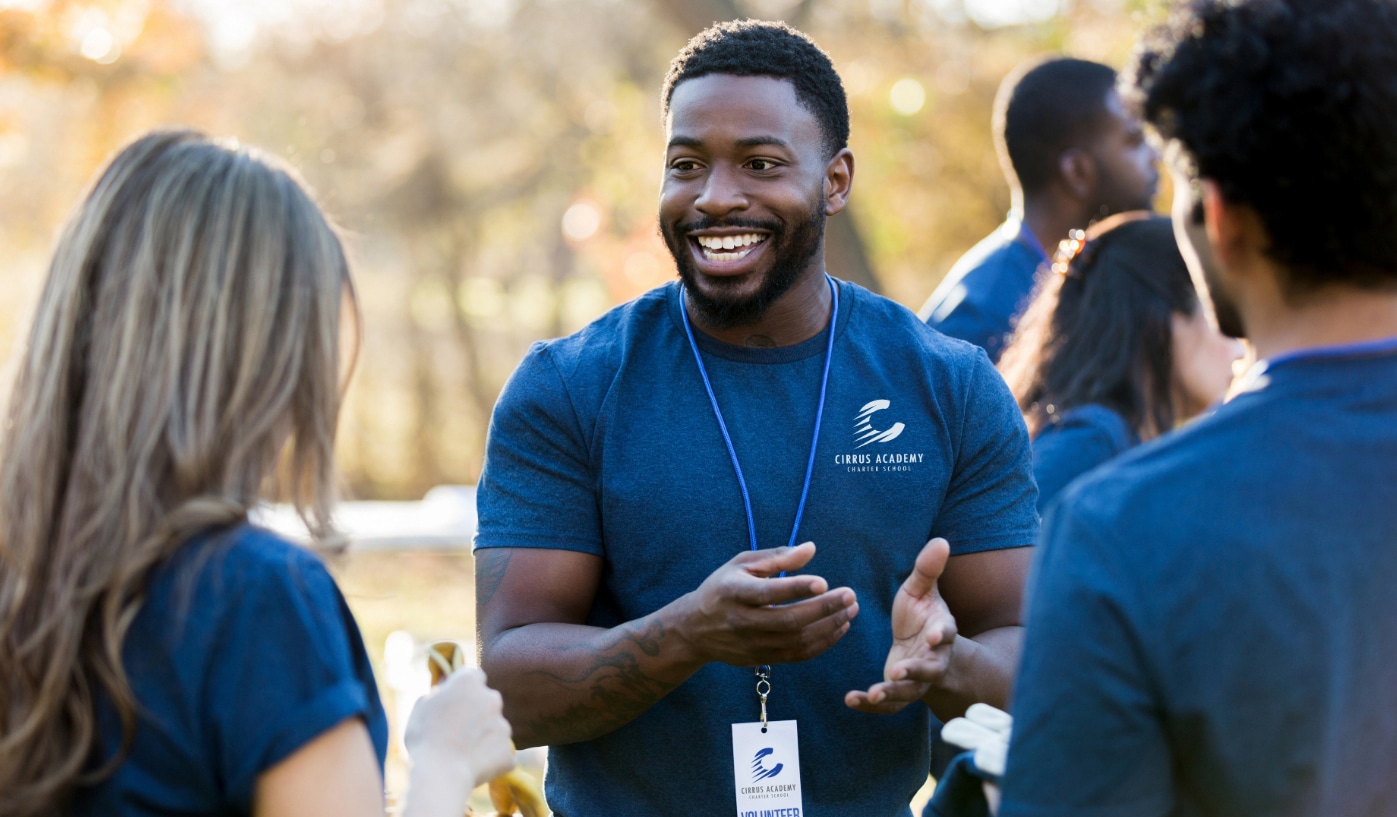 Man in blue t-shirt and name badge smiles, chatting with two people outdoors at an event with others and trees in the background.