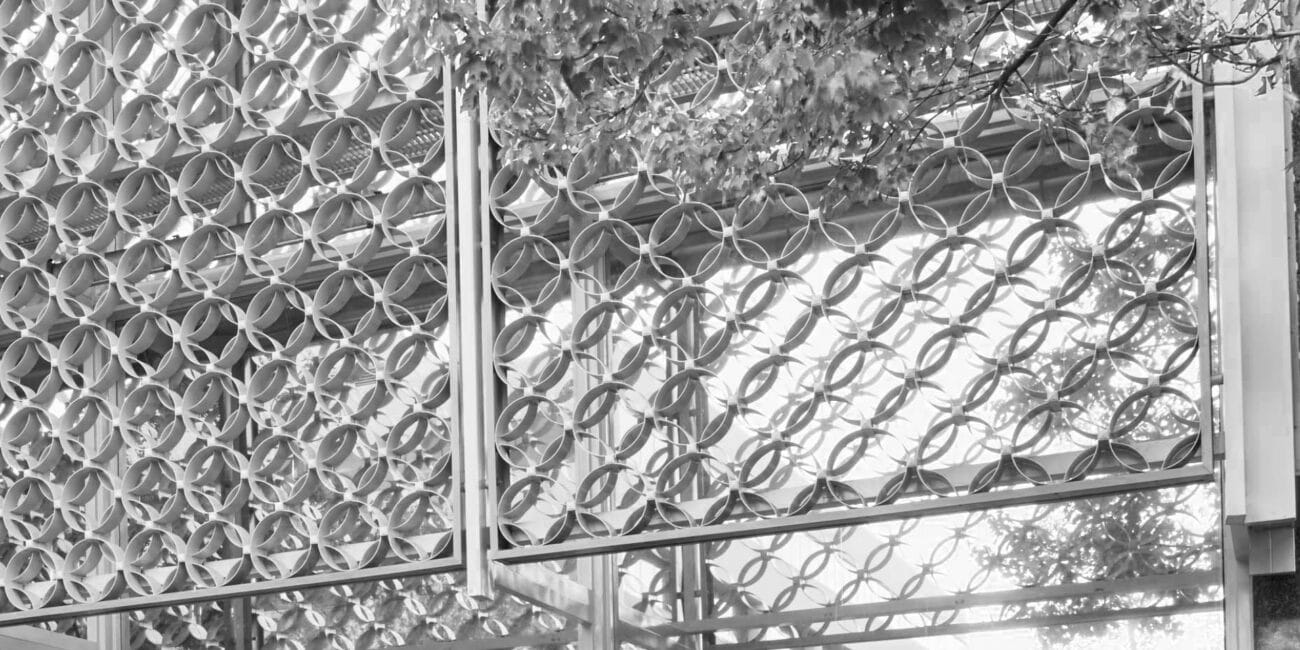 Geometric metal screen with circular pattern on building facade, partly covered by leafy branches; glass windows reflect light below.